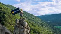A WVU student on top of a mountain top holding a flag with the flying WV and the words "Let's go."
