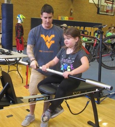 Man in WVU t-shirt helping young girl operate a rowing machine. 