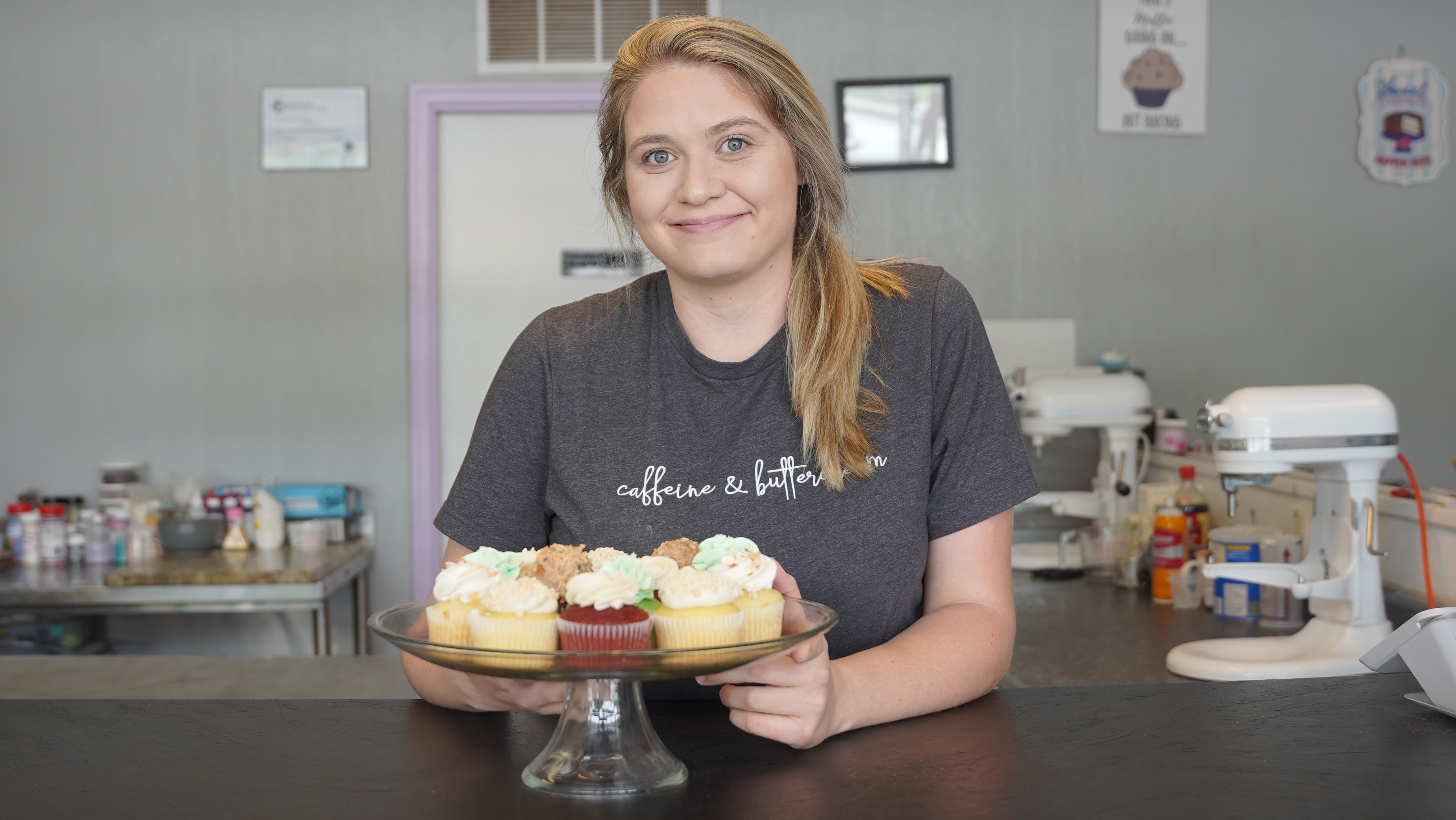 a photograph of Samantha Tichnell, smiling and resting her arms on a surface.  A plate of cupcakes is on the surface in front of her